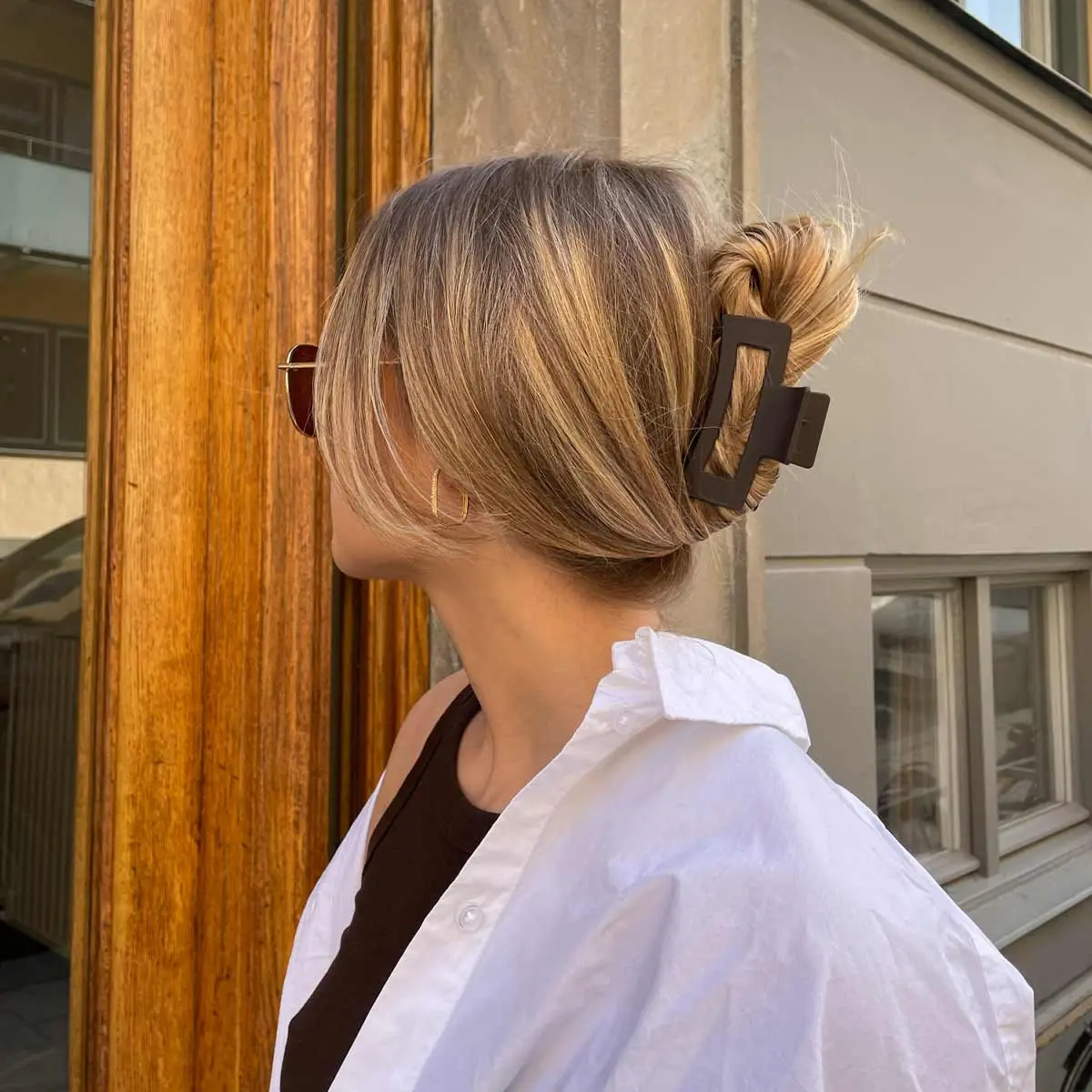 Braune Haarspange im Dutt – gestylt in blondem Haar, Modell mit Sonnenbrille und weißem Hemd vor einem Eingang.